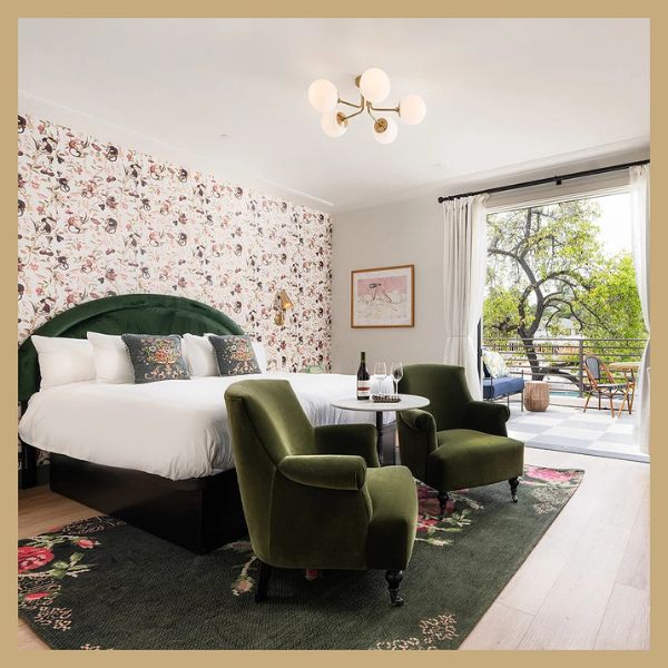 A wide view of a guest room with a bed, two green chairs, a small table, and a balcony door leading outside.