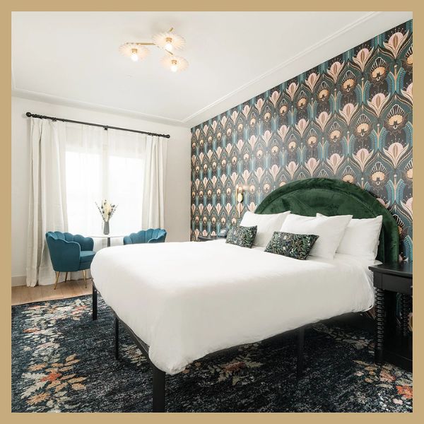 A wide view of a guest room with a large bed, floral wallpaper, and blue chairs by the window.