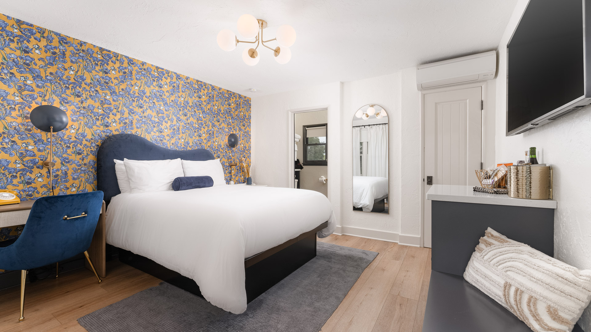 A wide view of a guest room with a blue and yellow wallpaper accent wall, a white bed with blue accents, a desk area with a blue chair, and a flat-screen TV mounted on the wall.