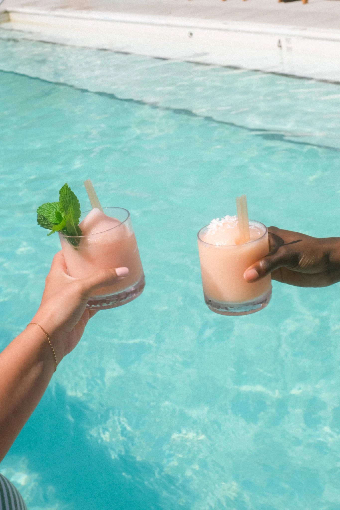 two hands holding glasses with drinks in front of a pool