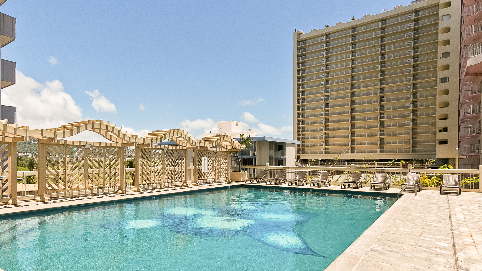 A hotel pool with lounge chairs and a pergola, surrounded by a high-rise building.