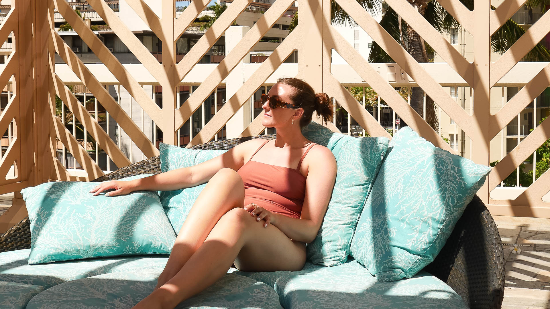 A woman in a pink swimsuit sits on a wicker sofa with blue pillows in an outdoor seating area with a wooden lattice wall and city view.