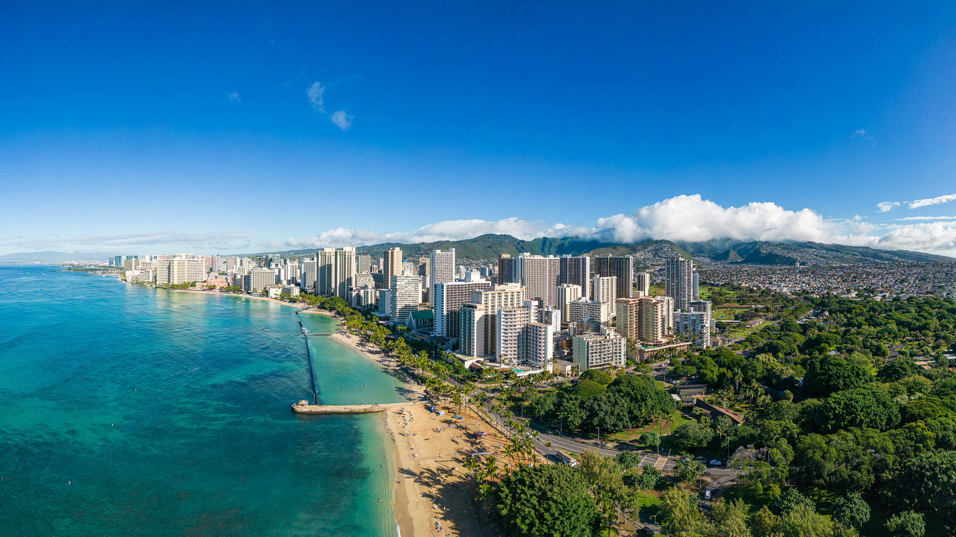 Aerial view of a beachfront city with high-rise buildings, trees, and a mountain range in the background.