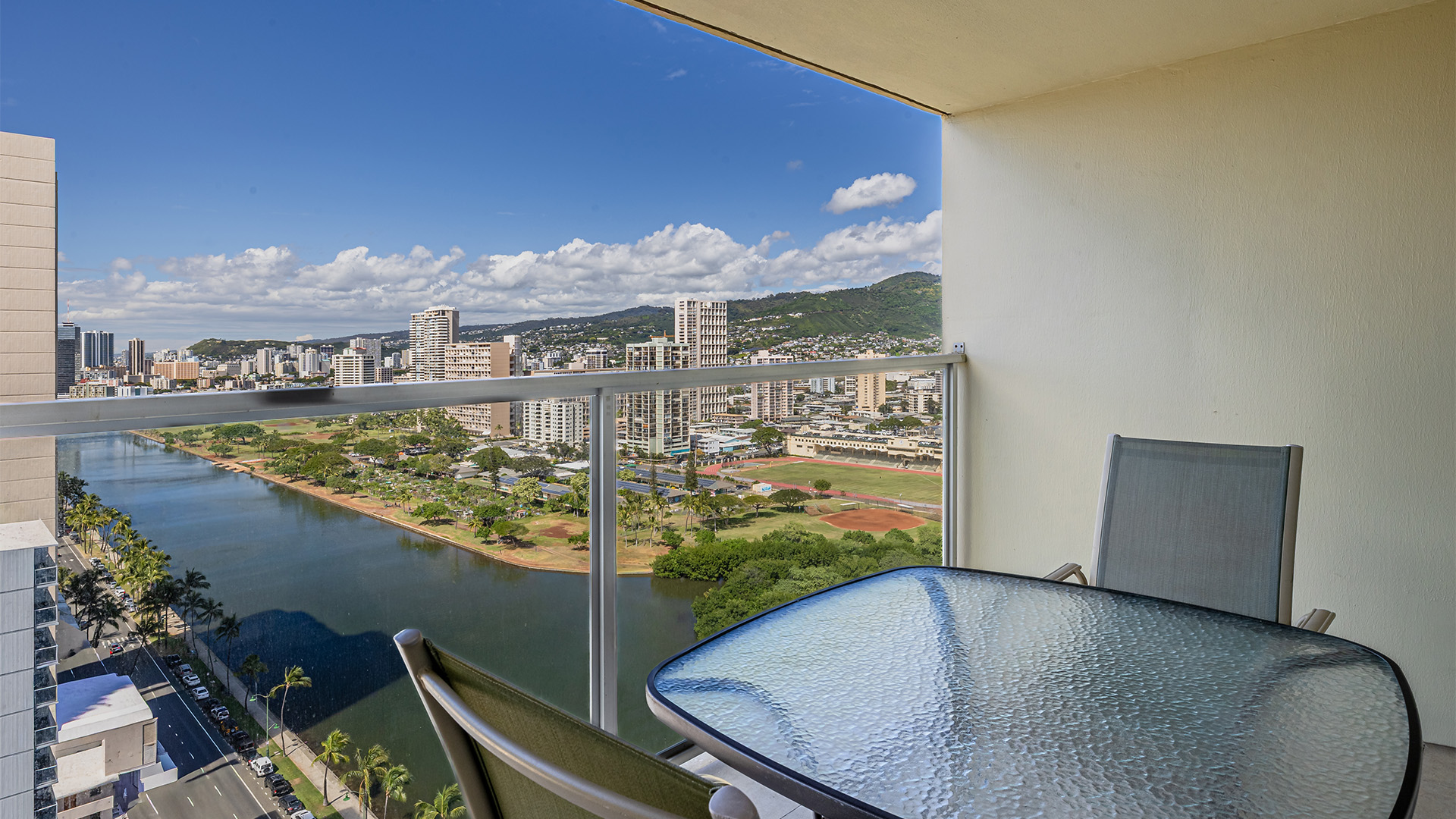 A balcony with a glass table and chairs overlooking a city and a river.