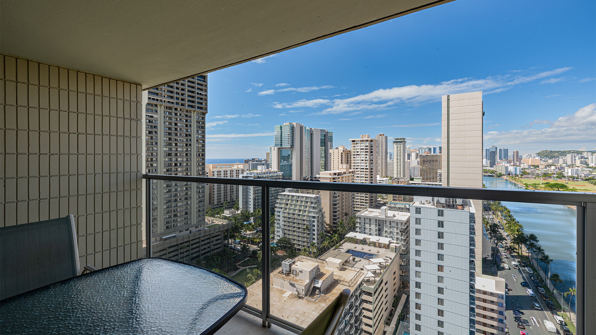 A balcony with a glass table and chair overlooking a cityscape with a body of water and blue sky.