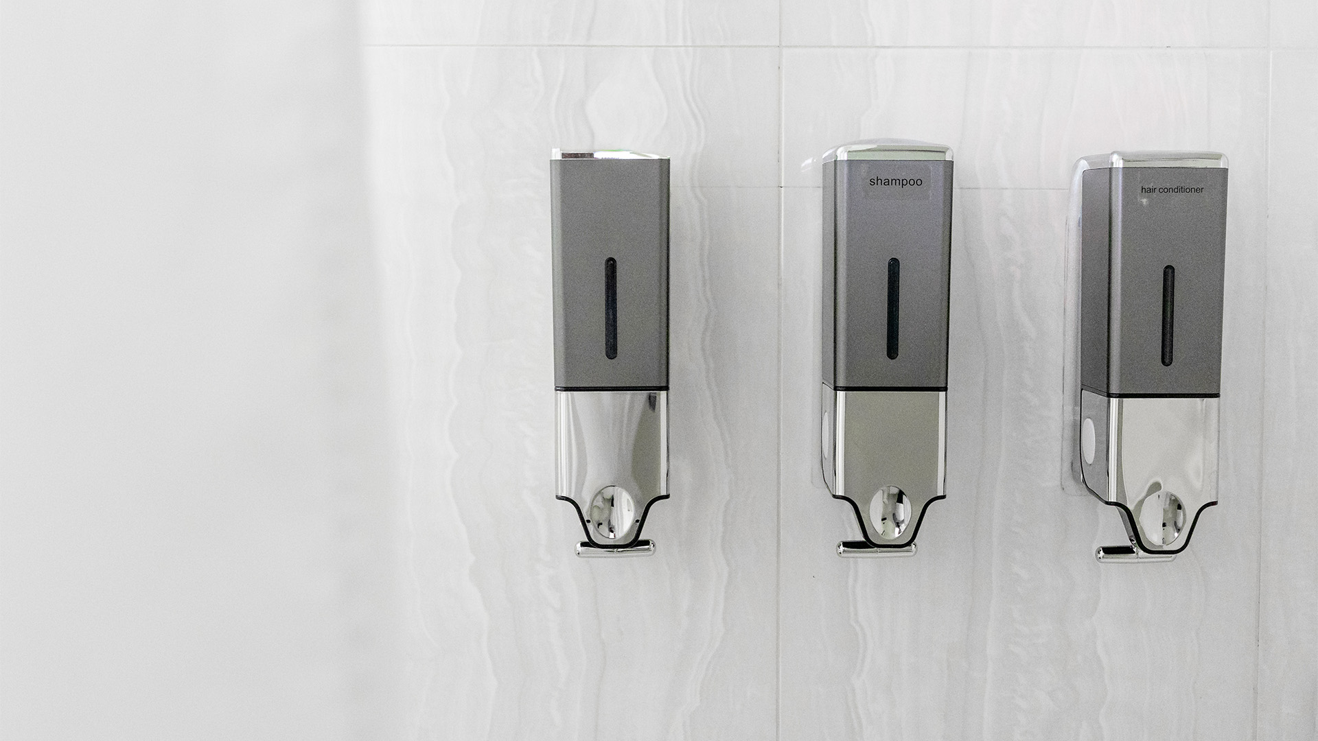 Three wall-mounted dispensers for soap, shampoo, and conditioner in a bathroom.