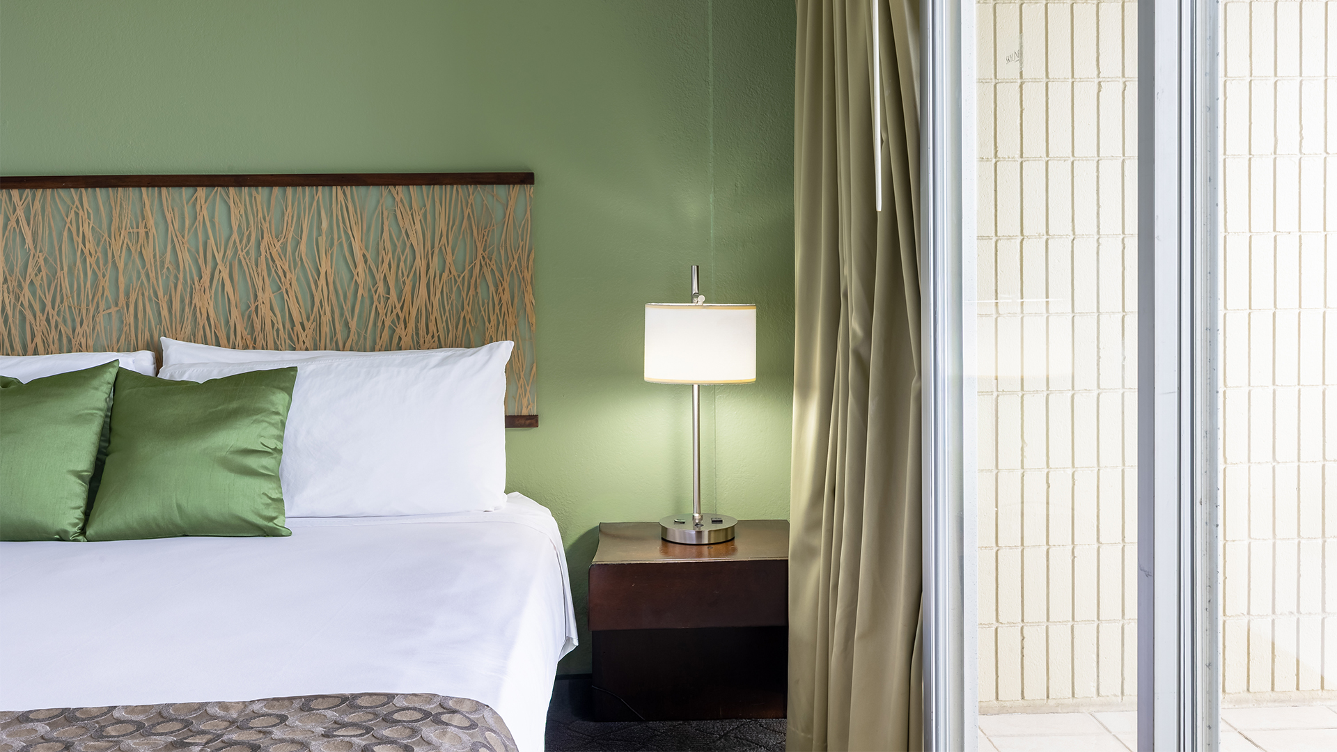A neatly made hotel room with a green wall, wooden headboard, and a reading lamp beside the bed.