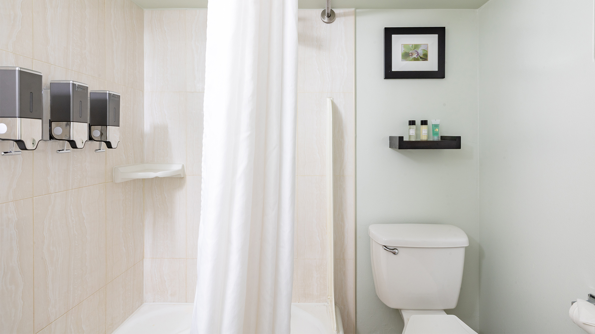 A standard hotel bathroom with tiled walls, a shower curtain, toilet, soap dispensers, and a shelf with toiletries.