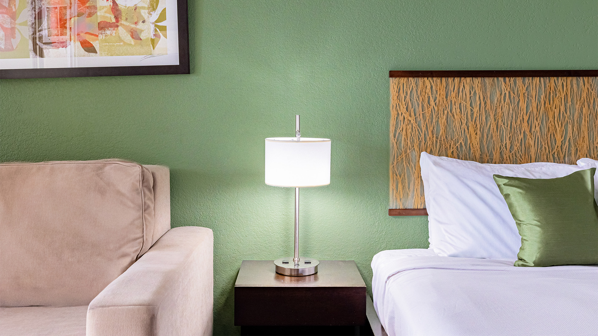 A hotel room with a green wall, a bed with white linens and a green pillow, a beige chair, and a lamp on a nightstand.