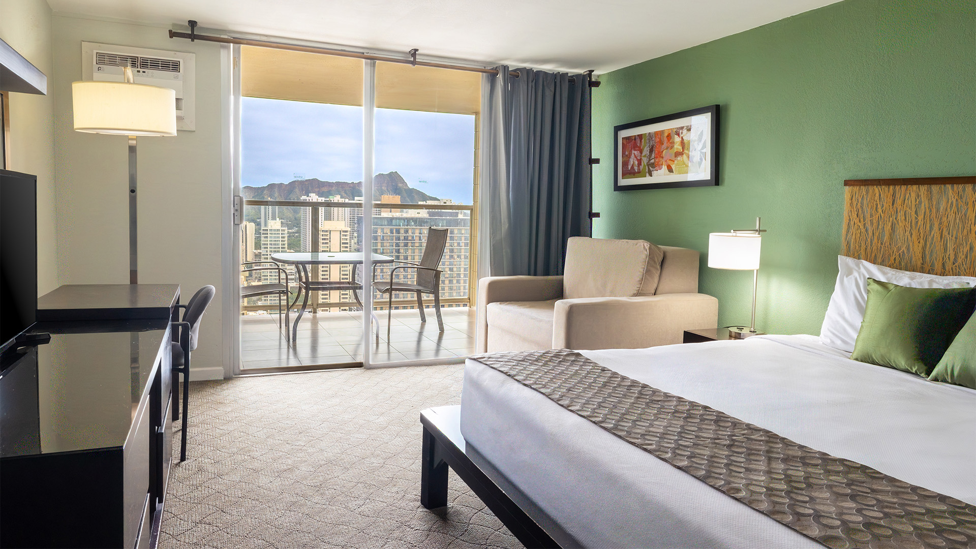 A hotel room with a bed, chair, desk, and balcony with a city view.