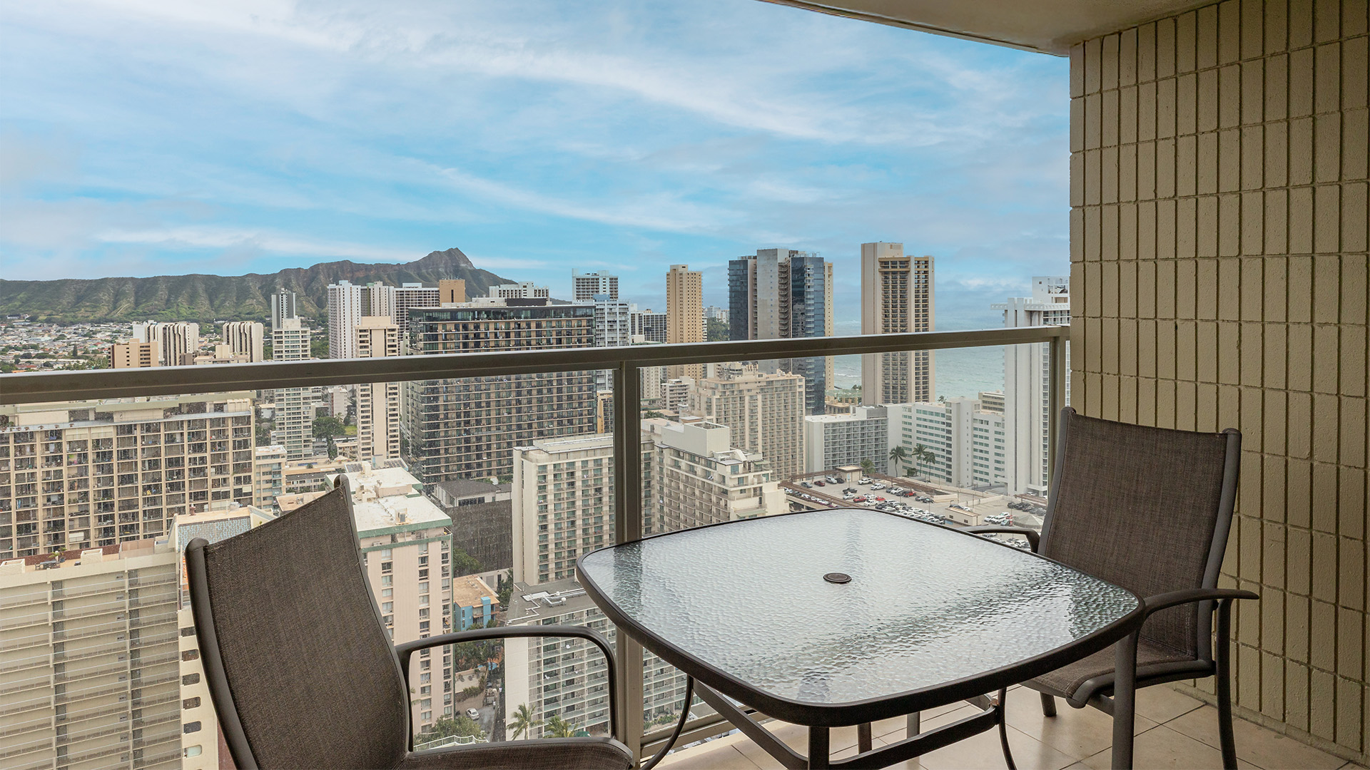 A balcony with a glass table and two chairs overlooking a cityscape with a mountain in the distance.