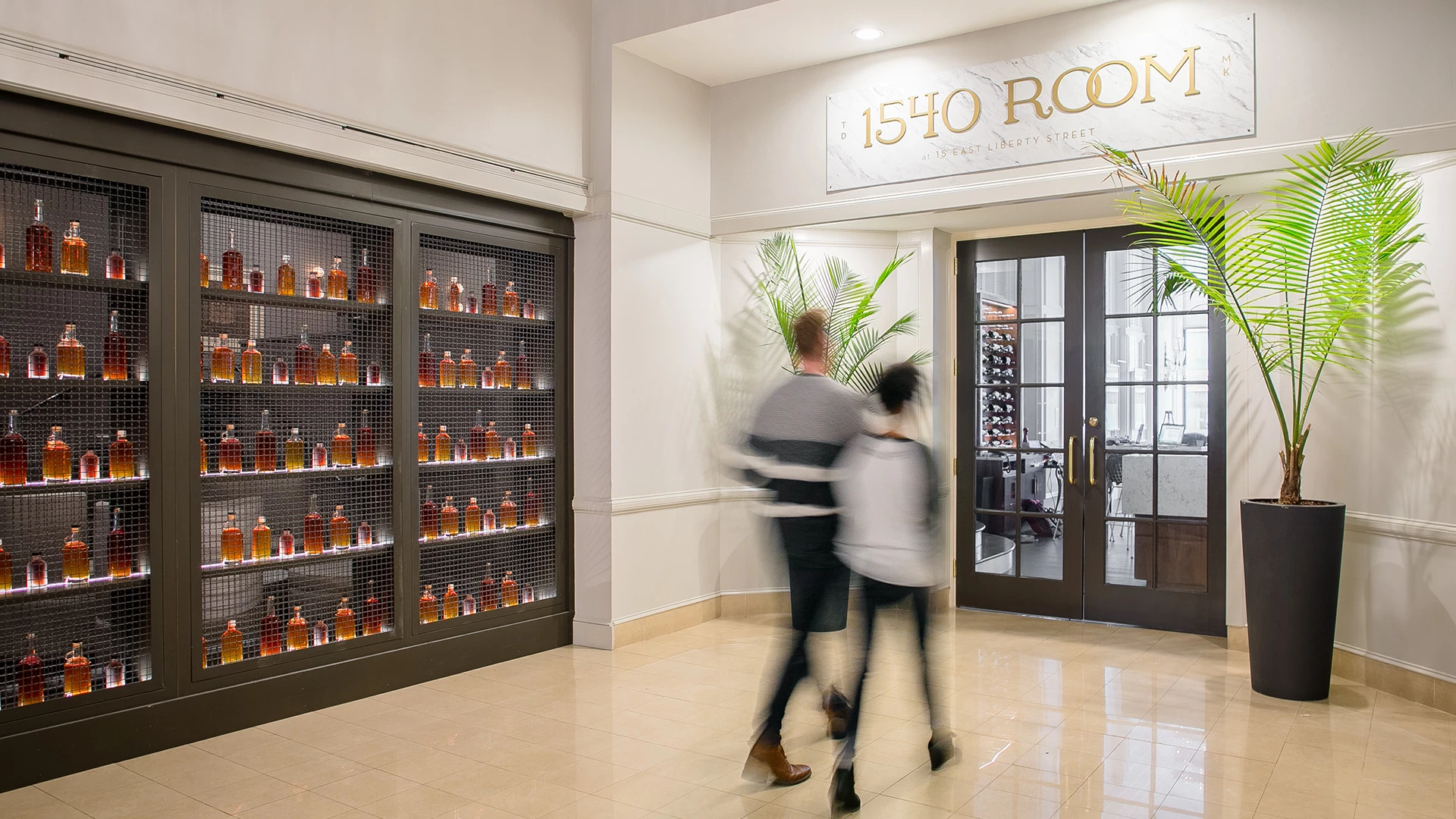 A lobby bar with a glass-enclosed wine storage, a tiled floor, and a potted plant, with two people walking by.