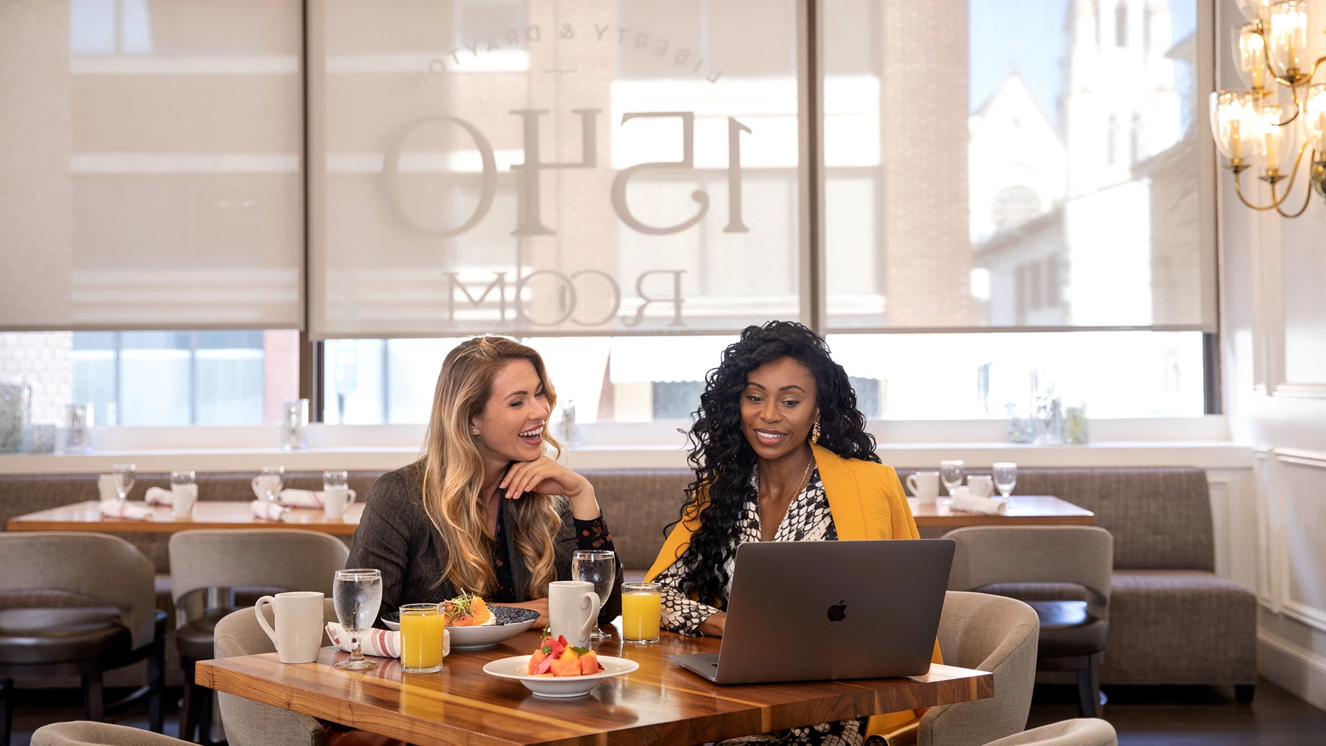 Two women sitting at a table in a dining room with a laptop and breakfast.