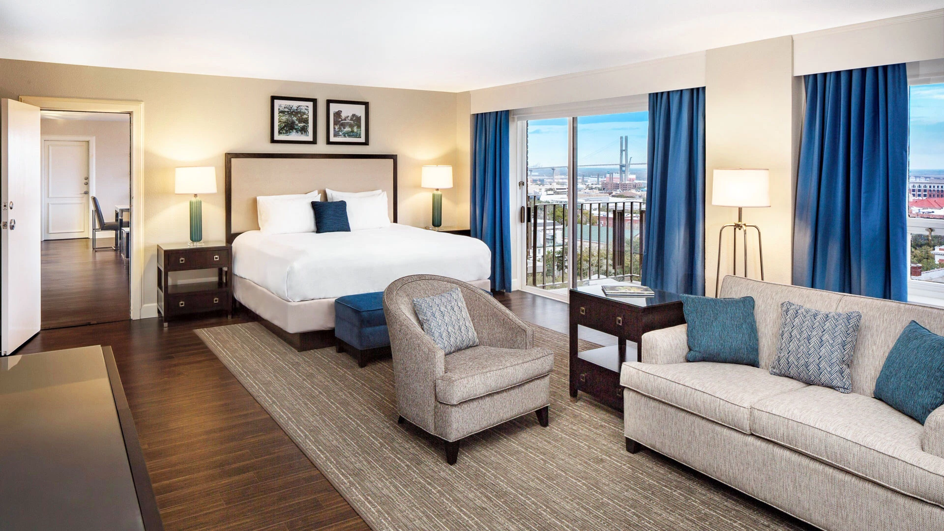 A hotel room with a large bed, sofa, chair, and balcony with a city view.