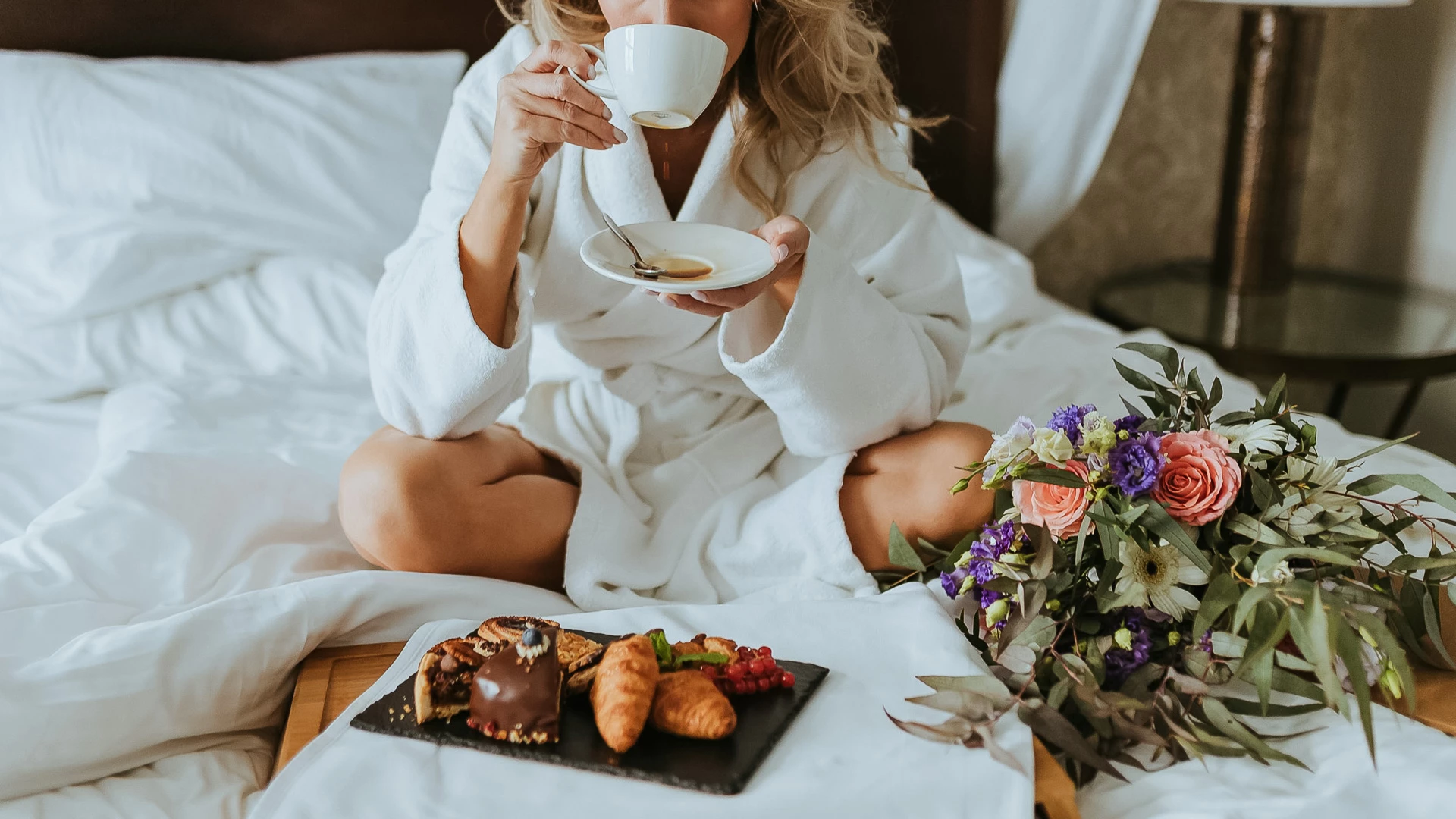 A woman in a white robe sits on a bed with white bedding, holding a cup of coffee and a saucer, with a breakfast tray and flowers nearby.