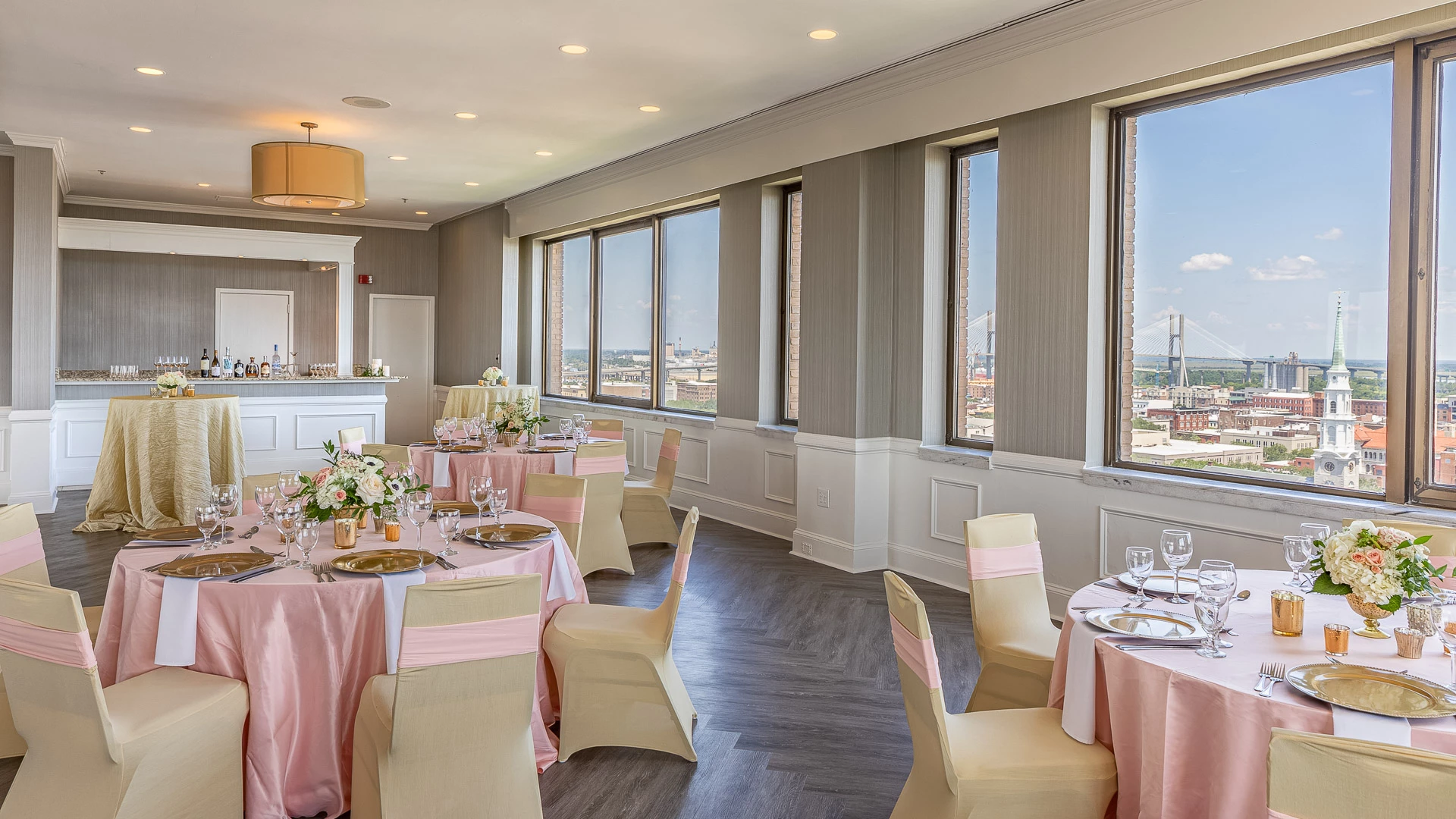A dining room with round tables set for a meal, a bar area in the background, and large windows offering a city view.
