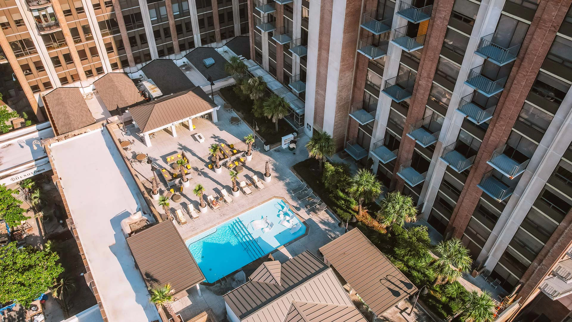An aerial view of a hotel pool area with a rectangular pool, lounge chairs, and buildings in the background.