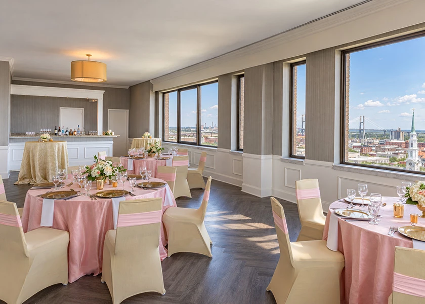 A dining room with round tables set for a meal, pink tablecloths, white chairs, and floral centerpieces, with a city view through large windows.