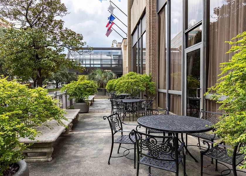 A small outdoor seating area with black metal tables and chairs, surrounded by plants and a brick building.