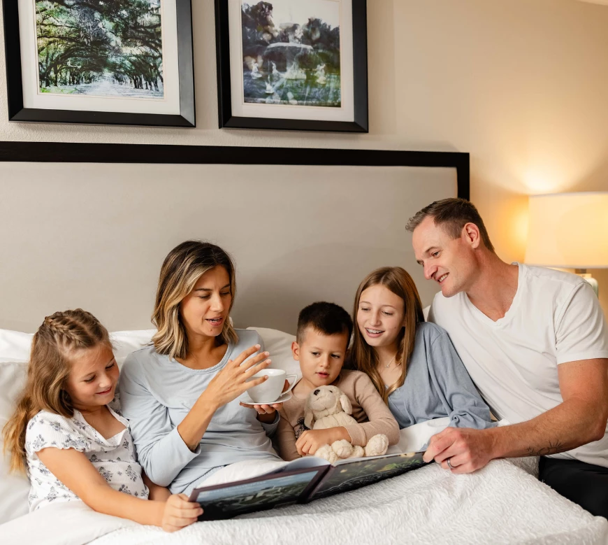 A family of five sits on a bed in a hotel room.