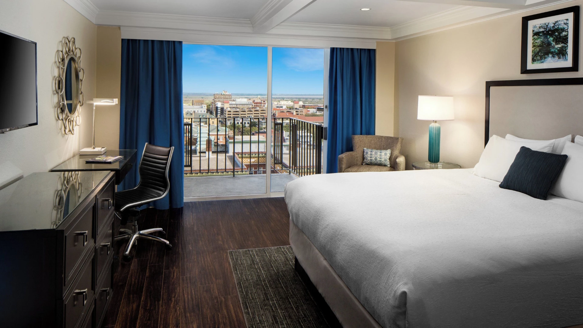 A hotel room with a large bed, desk, chair, and balcony with a city view.