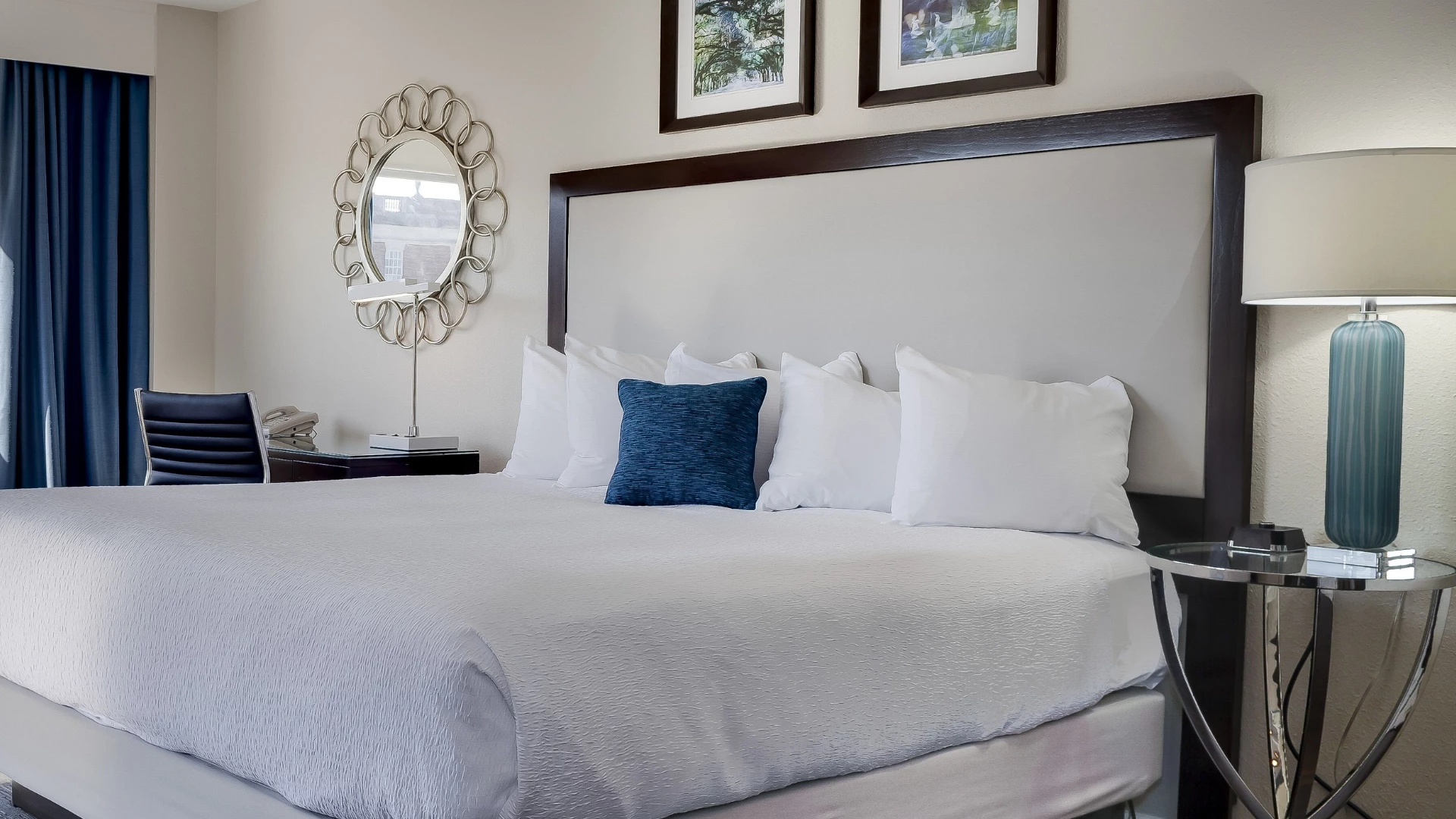 A hotel room with a large bed, nightstand, lamp, mirror, and desk.