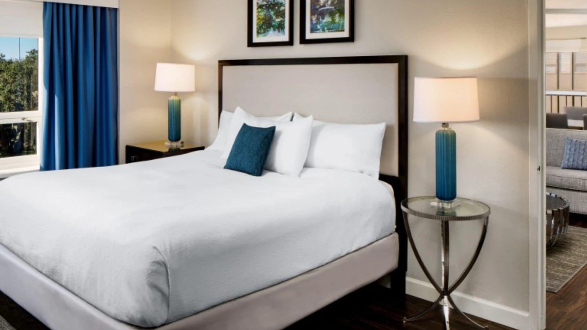 A neatly made king-sized bed with white linens and blue accent pillows, flanked by nightstands with lamps, in a room with a window and artwork.