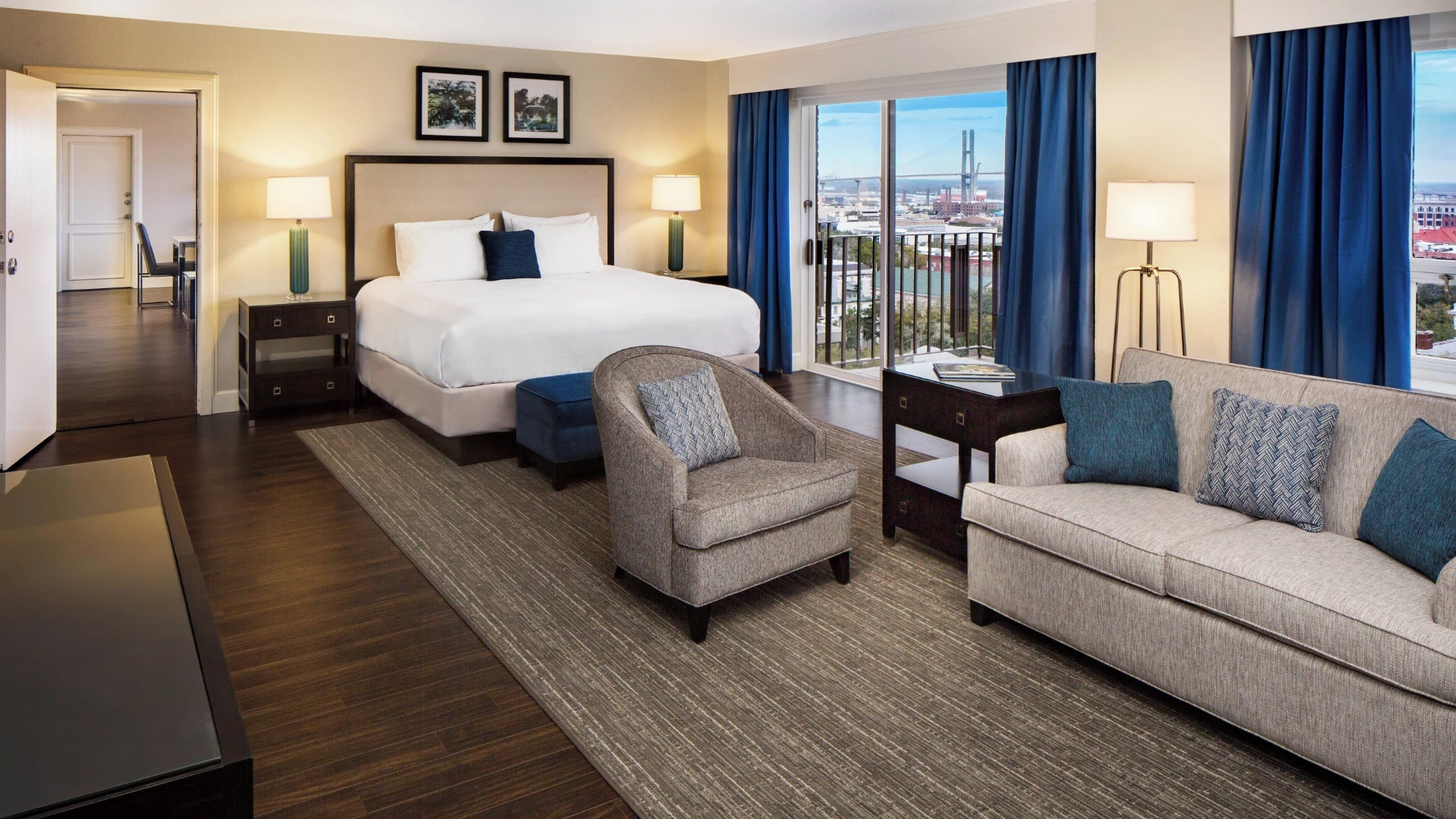 A wide view of a hotel room with a bed, sofa, armchair, and balcony with a city view.
