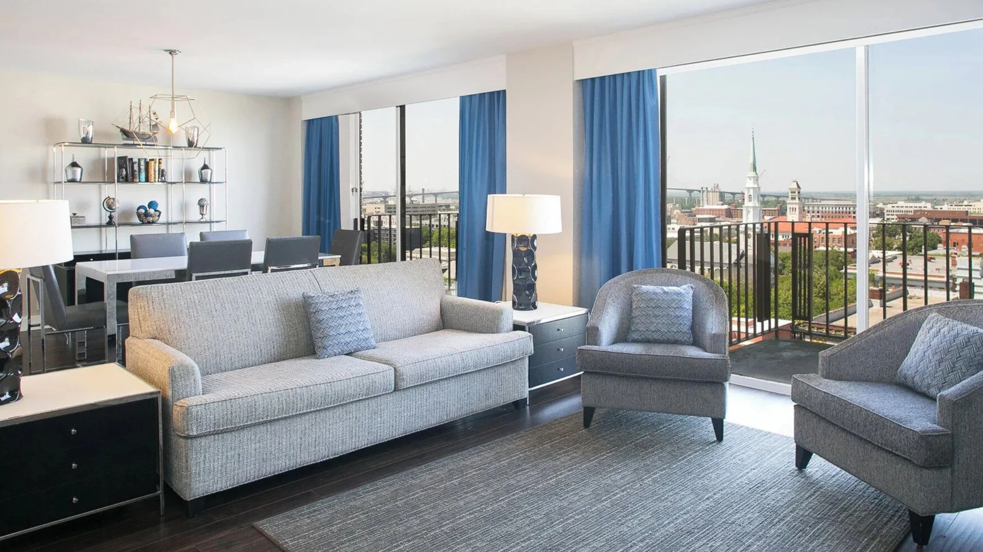 A wide view of a suite with a sofa, armchairs, a dining table, and a balcony with a city view.