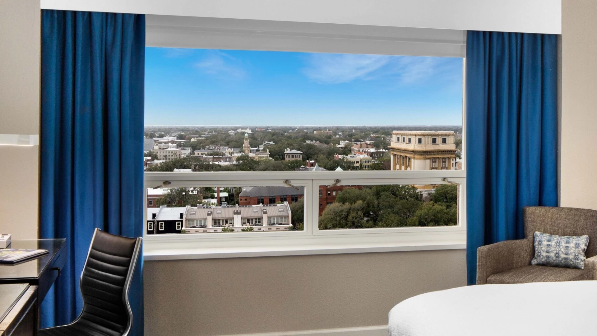 A hotel room with a large window showing a city view, a desk, chair, and bed.