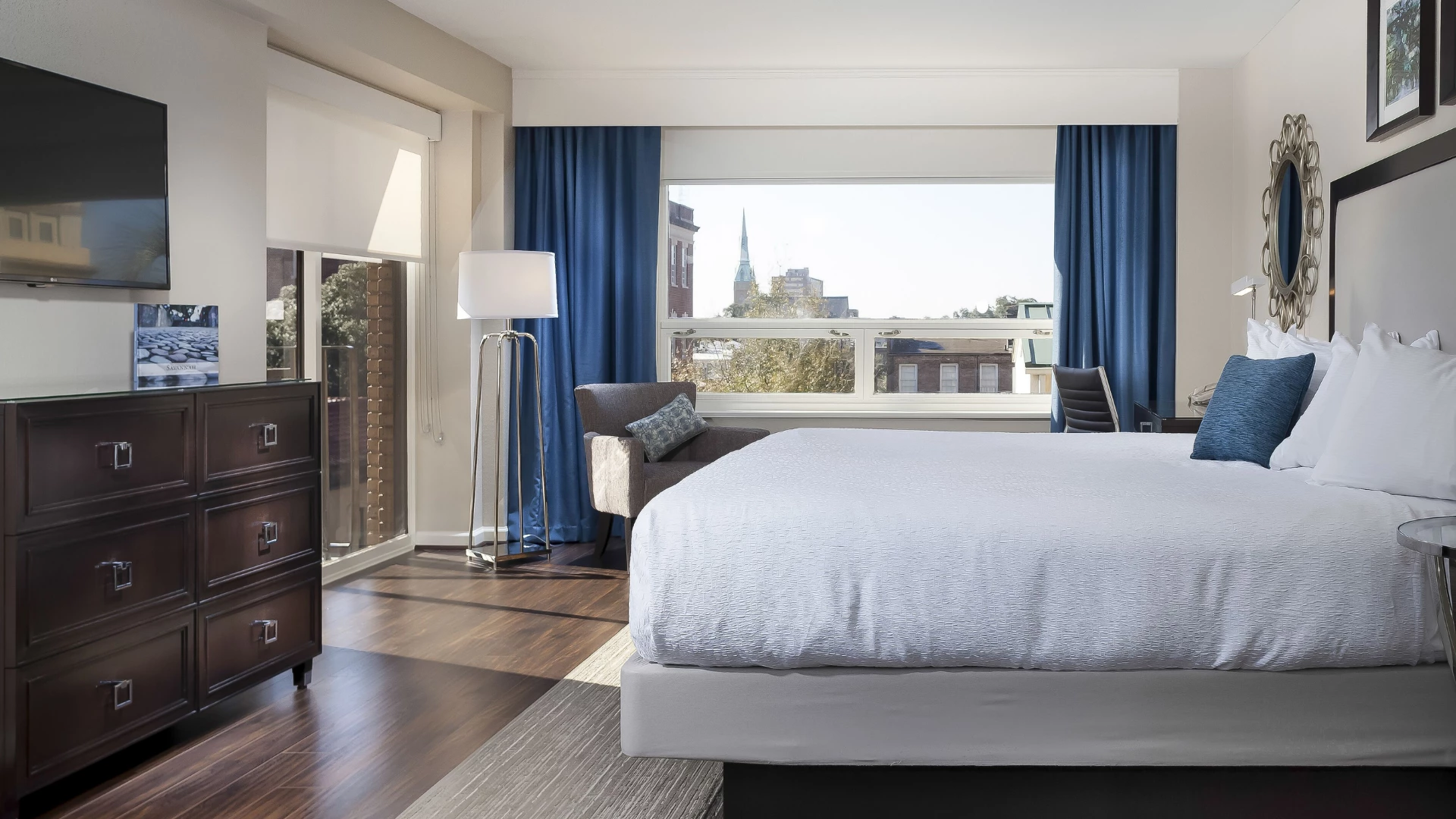 A large hotel room with a king-sized bed, dark wood dresser, armchair, and city view.