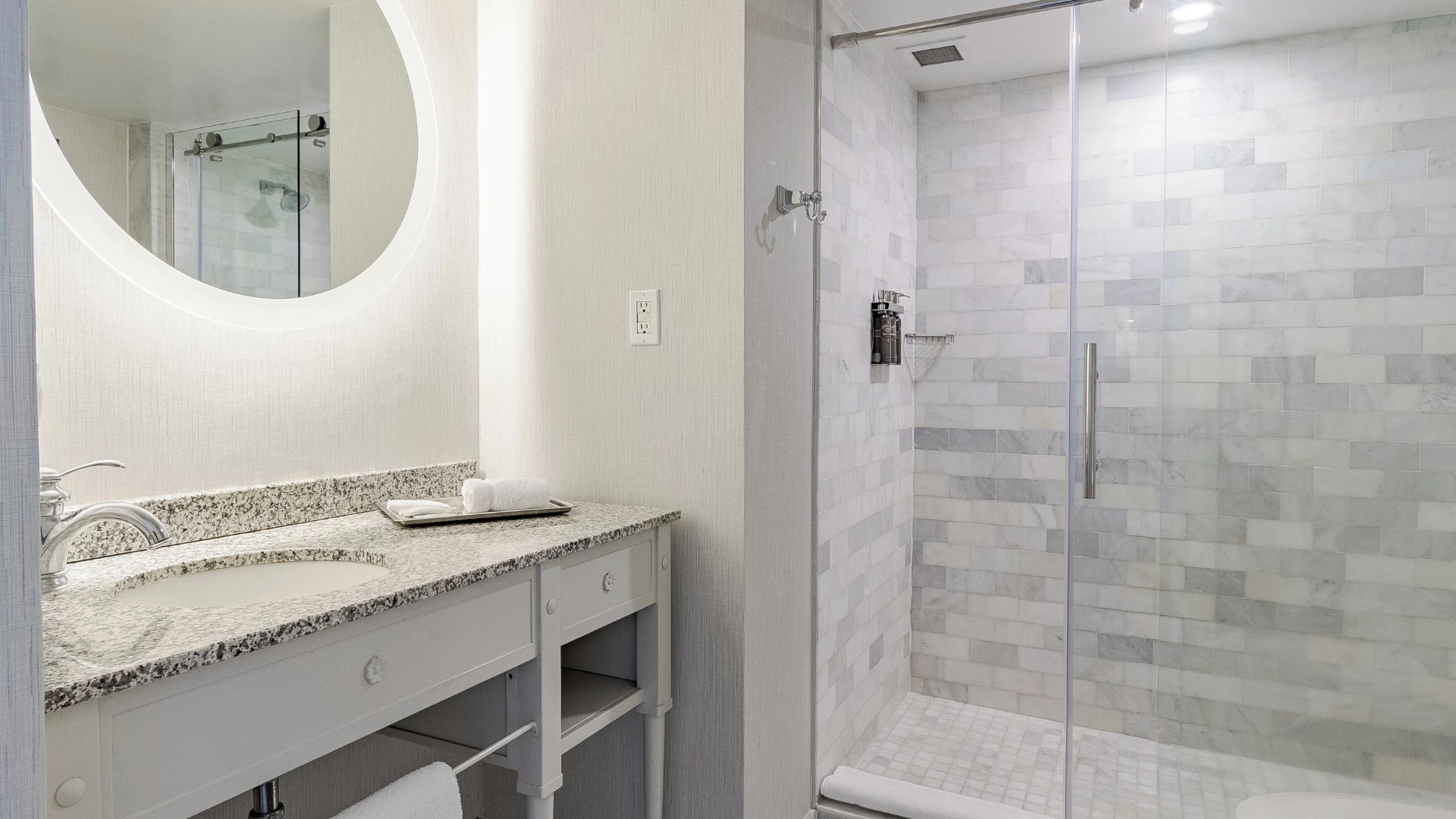 A clean bathroom with a granite countertop, glass shower, and round mirror