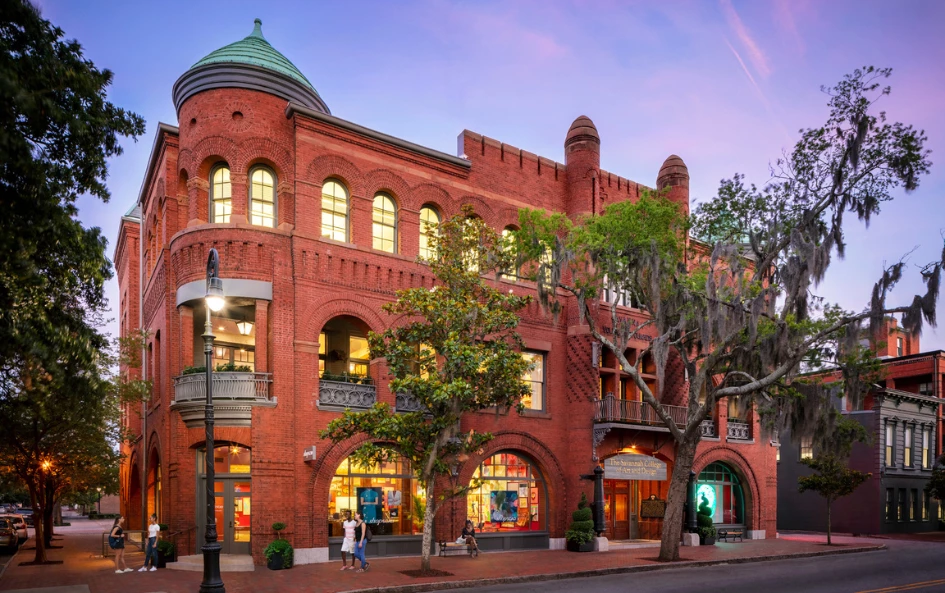 A red brick building with arched windows and a green dome sits on a street corner with people walking by.