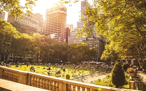 A sunny view of a park with people sitting on the grass and a city skyline in the background, seen from a balcony with a railing.