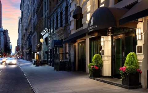 A city street with a hotel entrance featuring a black awning with white lettering, glass doors, and potted plants.
