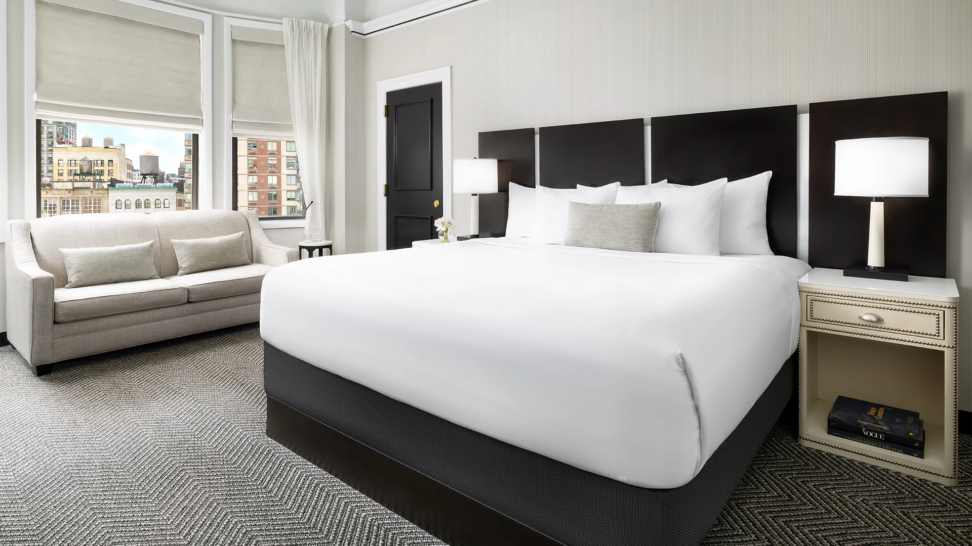 A wide view of a hotel room with a large bed, nightstand, sofa, and city view.