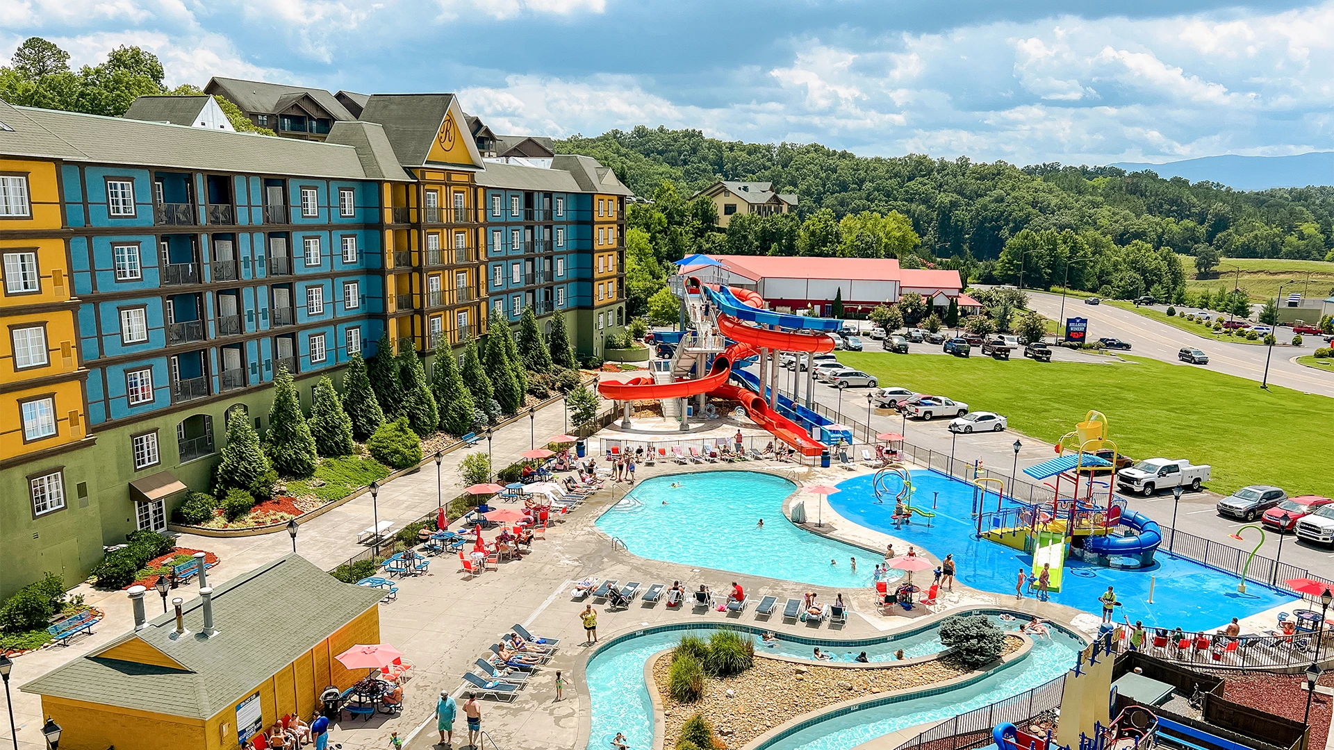 An aerial view of a hotel with a large pool area featuring water slides, lounge chairs, and a parking lot.