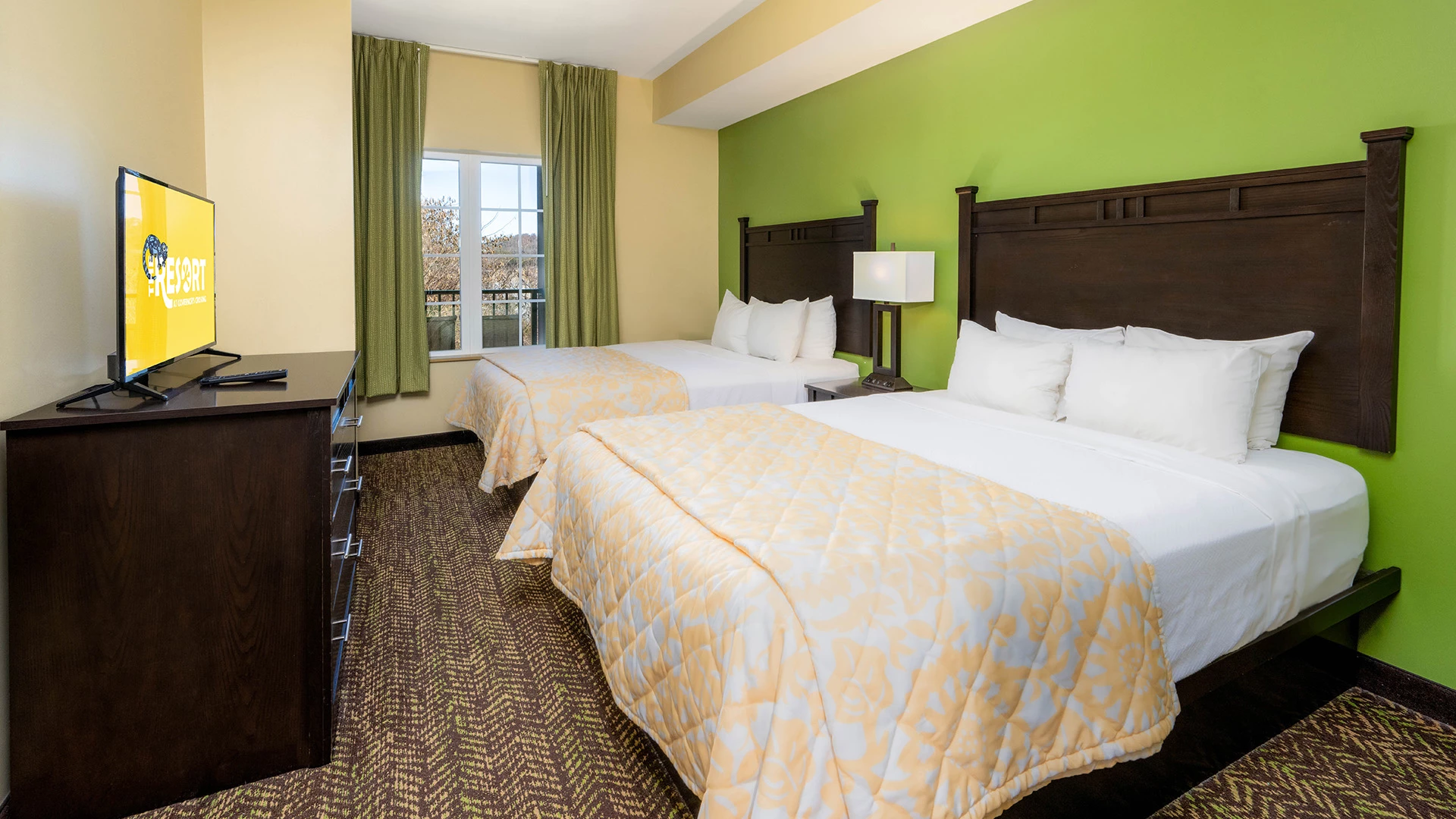 A hotel room with two twin beds, a dresser with a flat-screen TV, and a window with green curtains.