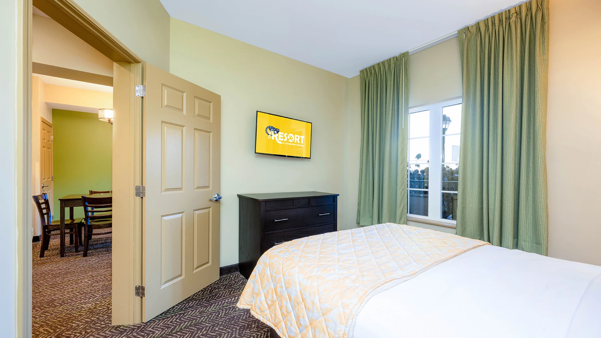 A wide view of a hotel room with a bed, TV, window, door, and dresser.