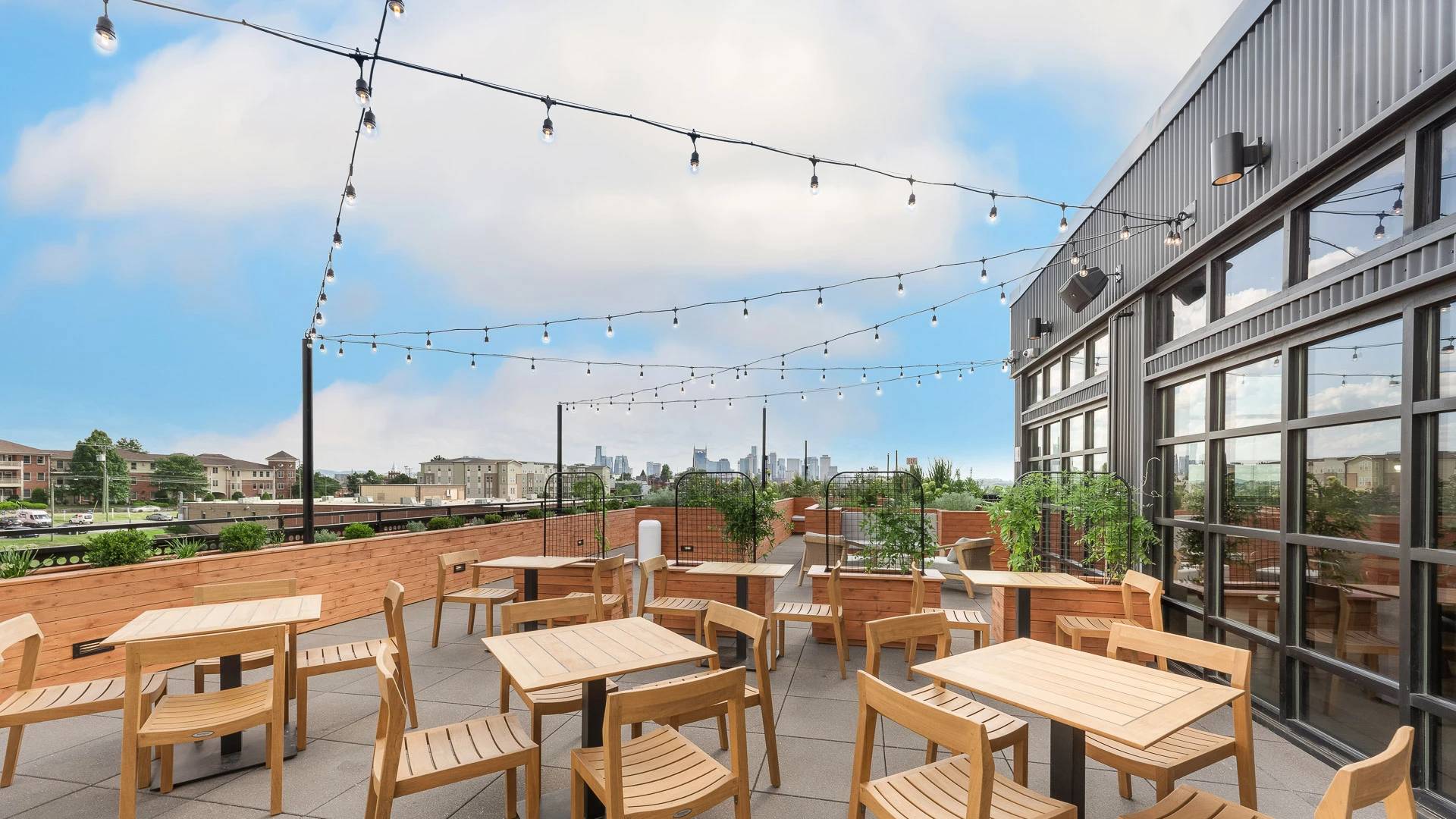 A rooftop outdoor seating area with wooden tables and chairs, string lights, and a city skyline view.