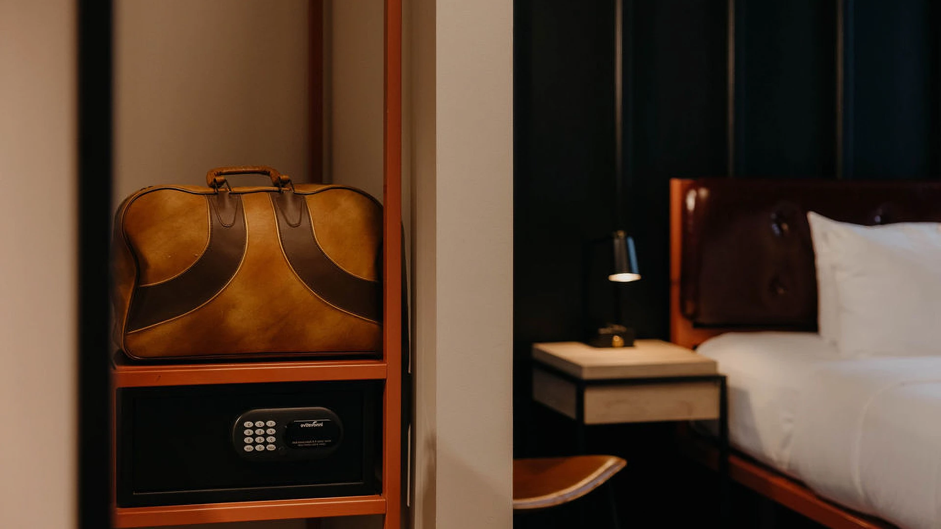 A hotel room with a leather suitcase on a shelf above a safe, a bed with white linens and a pillow, and a nightstand with a lamp.