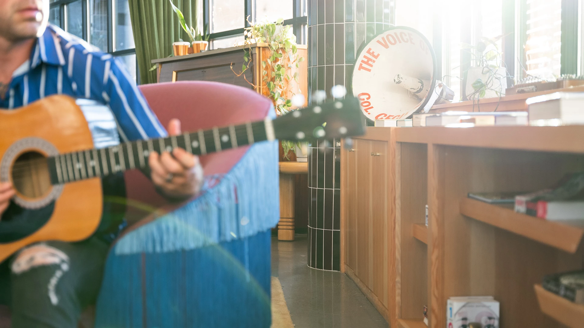 A man plays guitar in a lobby with a colorful chair and wooden shelves.