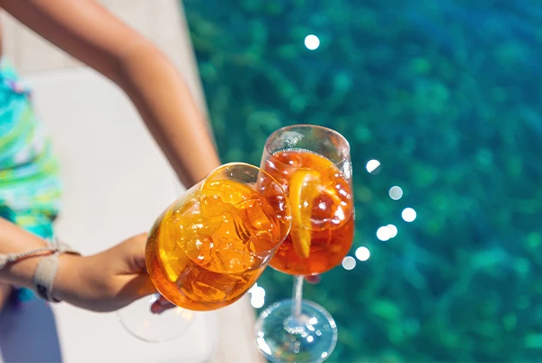 Two glasses of orange liquid with ice and lemon are held by a person next to a pool.