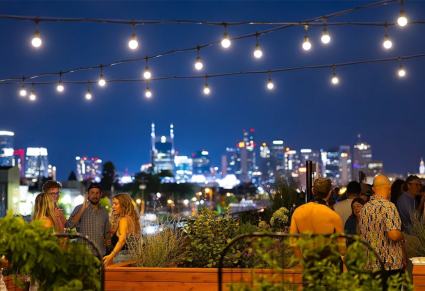 A group of people are gathered on a rooftop with string lights, plants, and a city skyline in the background.