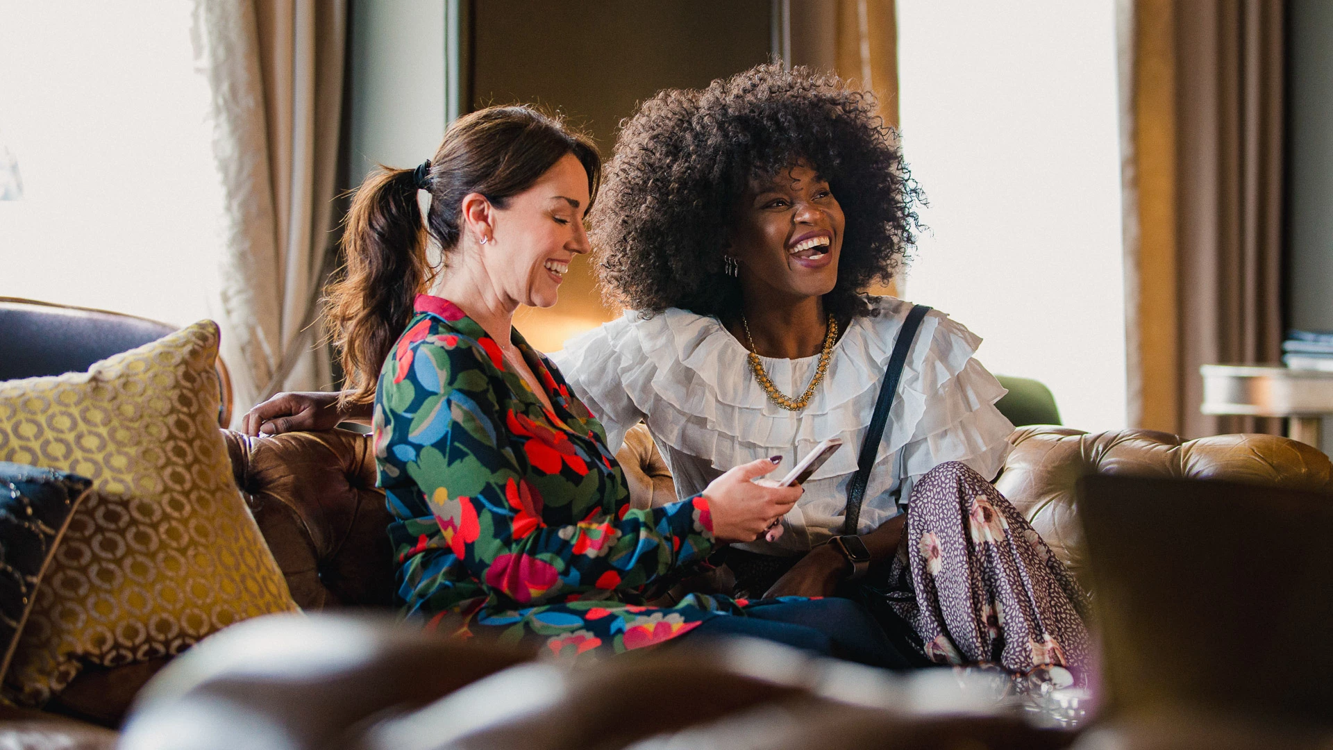 Two women sitting on a brown leather sofa in a lobby area with curtained windows.