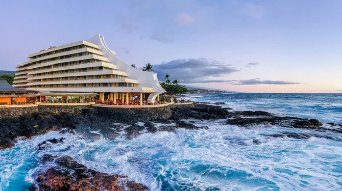 Royal Kona Resort hotel building overlooking ocean on a rocky shore with waves crashing on lava rocks below it 