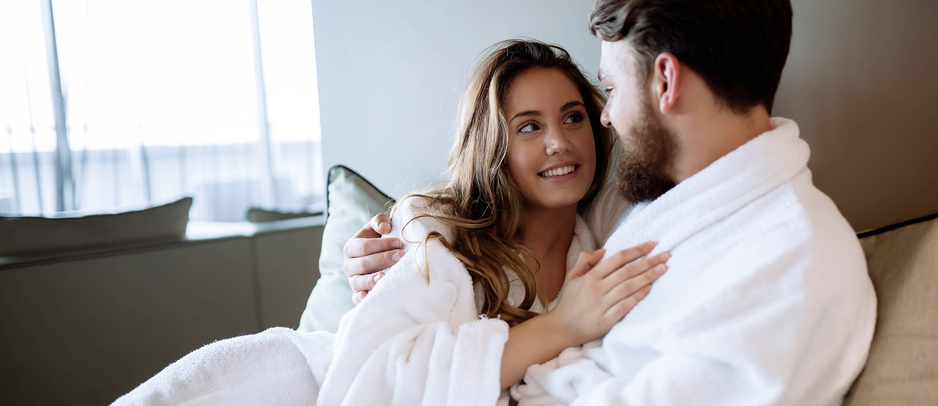 couple wearing hotel robes in bed and looking at each other