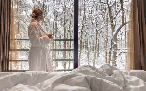 a woman in a white robe standing in front of a window with a view of snow covered trees