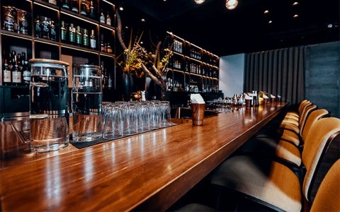 A dimly lit bar with a wooden counter, shelves of bottles, and stools.