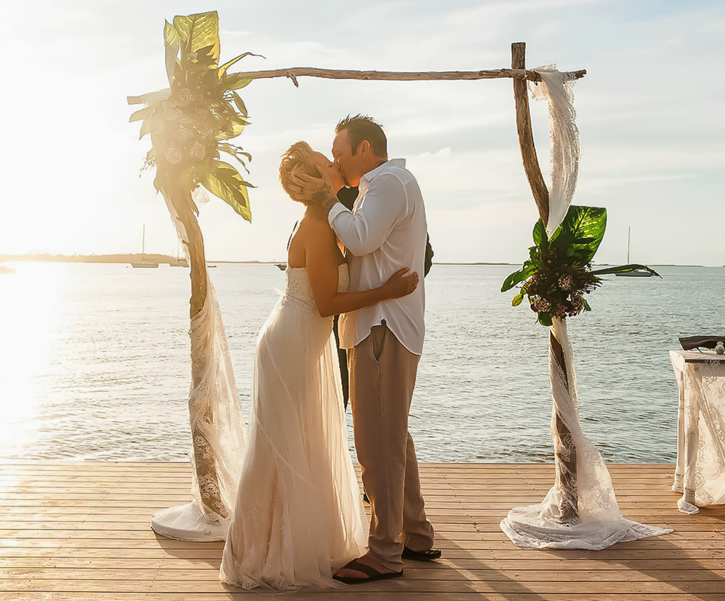 a man and woman kissing under a wooden arch with palm trees and water